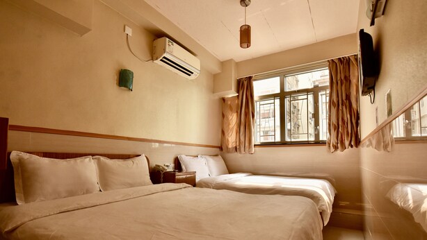 A hotel room with two beds, a window with curtains, an air conditioning unit, a bedside table, and a television mounted on the wall. - Wonderful Inn (Kowloon)