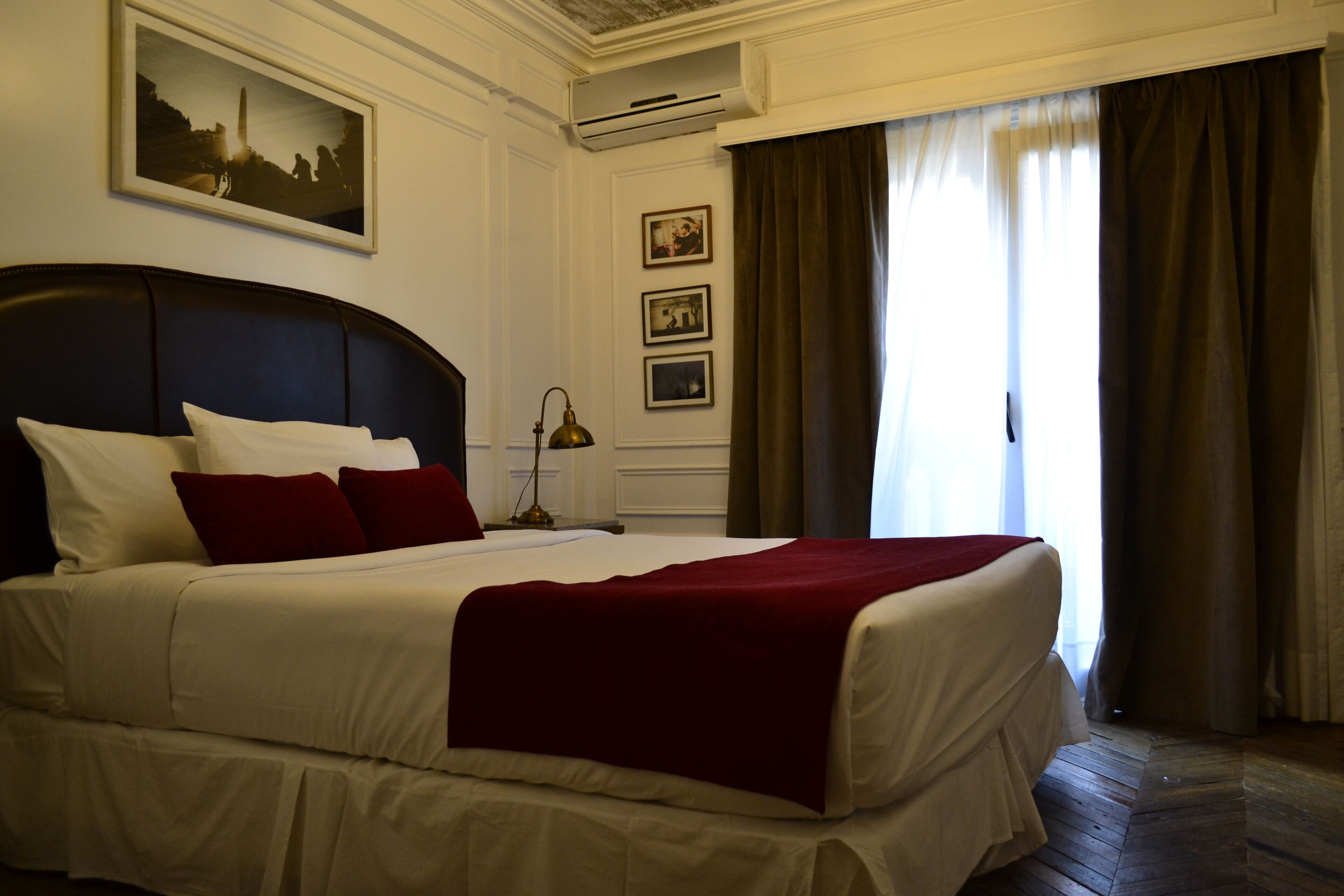 A neatly made bed with white linens and a red throw, a bedside lamp, and framed pictures on the wall.