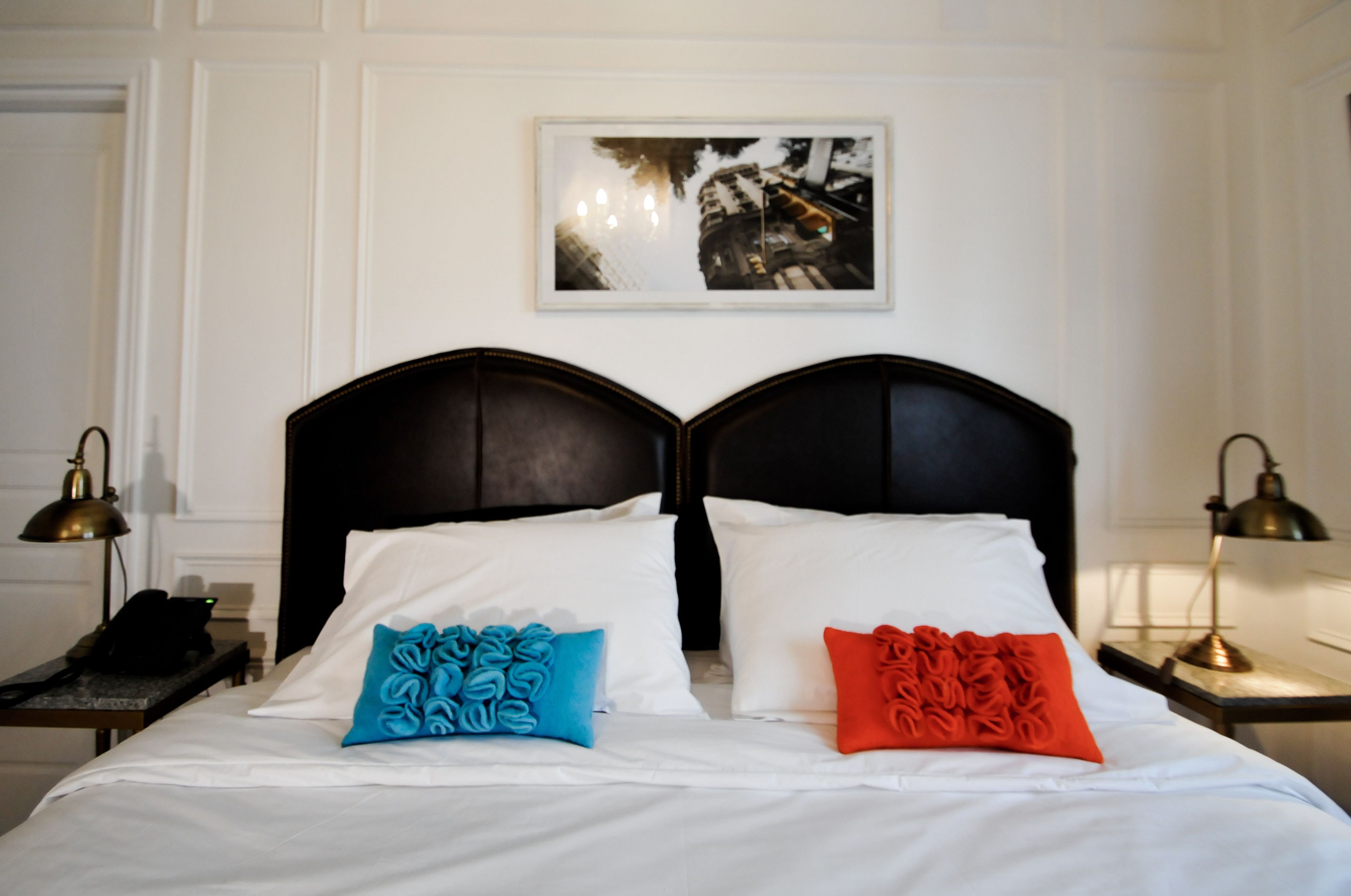 A double bed with white bedding and two pillows, one blue and one red, in a room with white walls and a framed picture above the bed.
