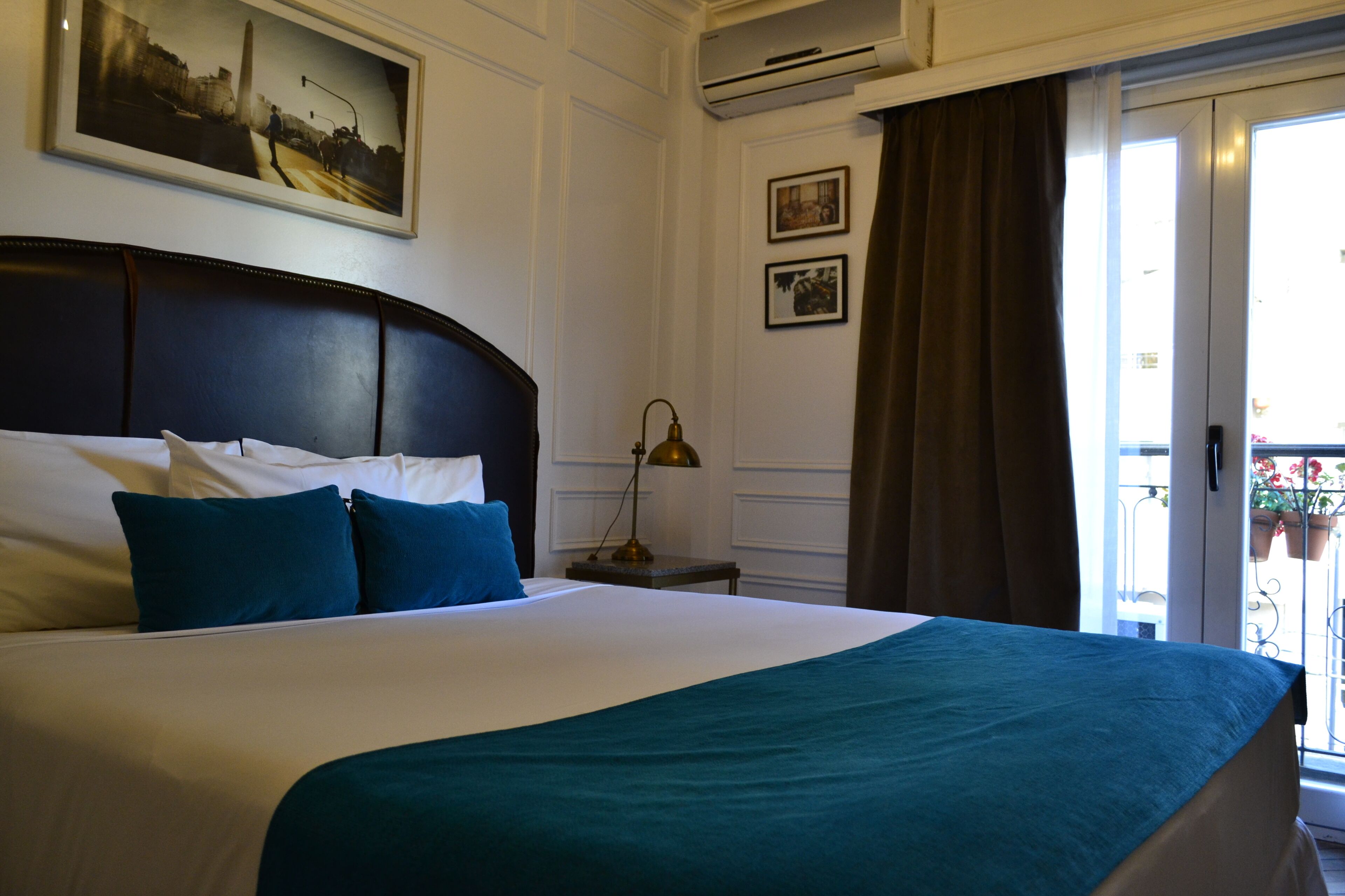 Habitación de hotel con una cama grande, una mesita de noche con lámpara, un cuadro en la pared y vistas al balcón exterior.