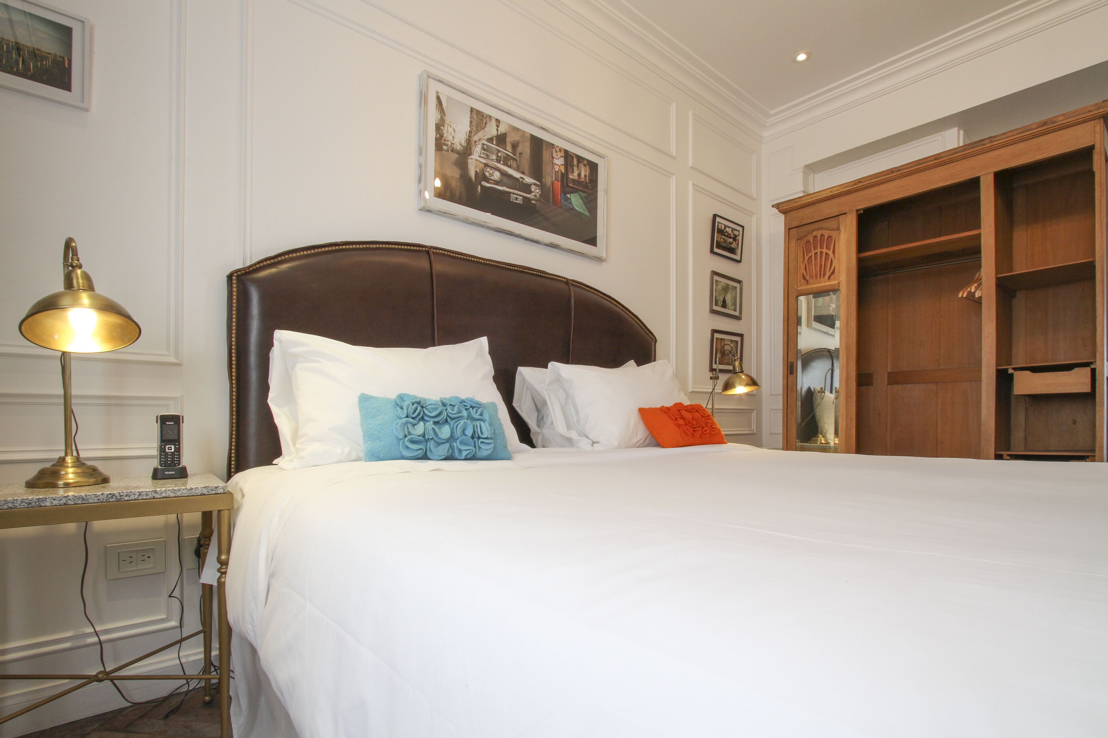A neatly made bed with white linens and decorative pillows, a bedside table with a lamp, and a wooden wardrobe.