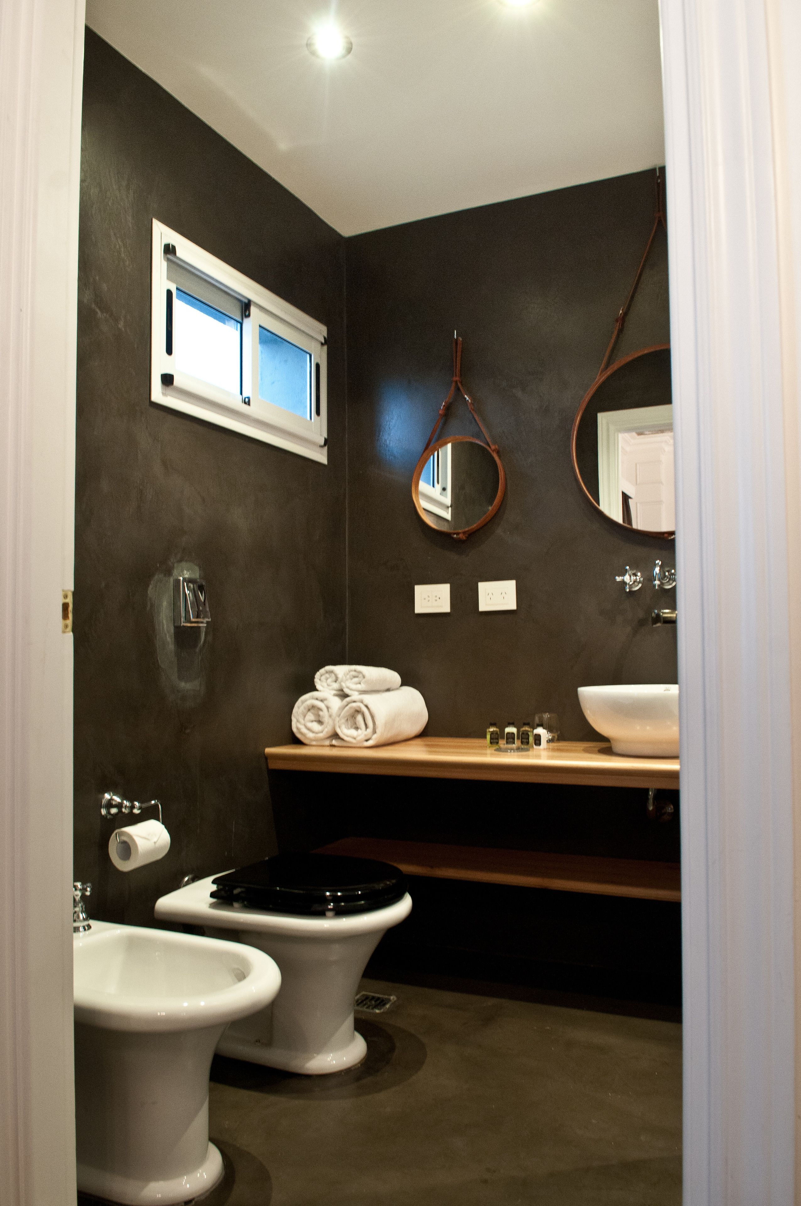 A bathroom with a dark wall, a window, a mirror, two toilets, a sink, and a towel rack.