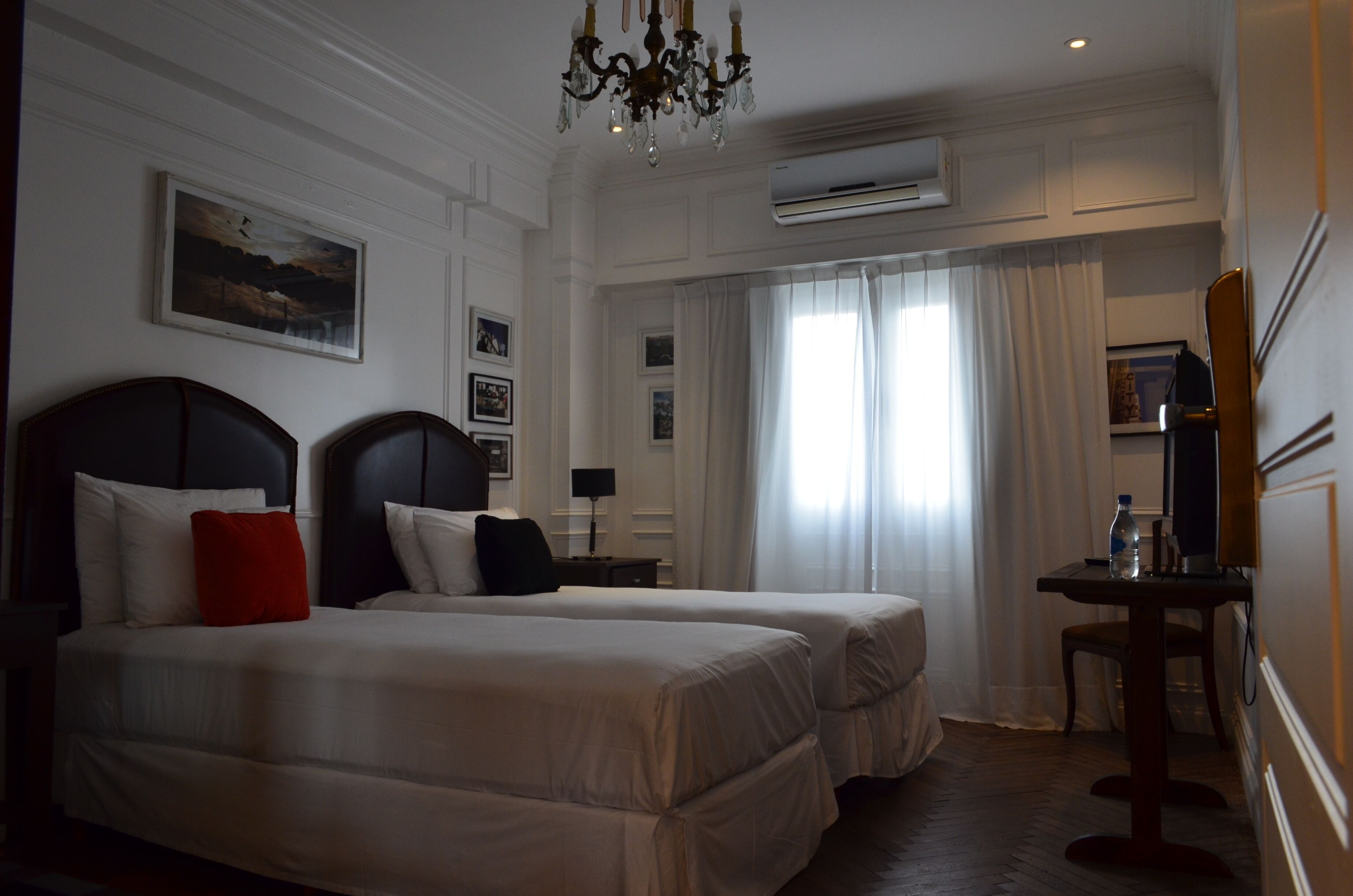 A hotel room with two beds, a desk, a chair, a chandelier, and an air conditioning unit.