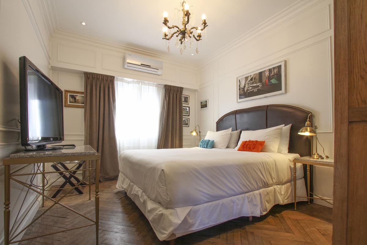 Una habitación de hotel con una cama grande, un televisor sobre un mueble bajo, un escritorio y una araña.