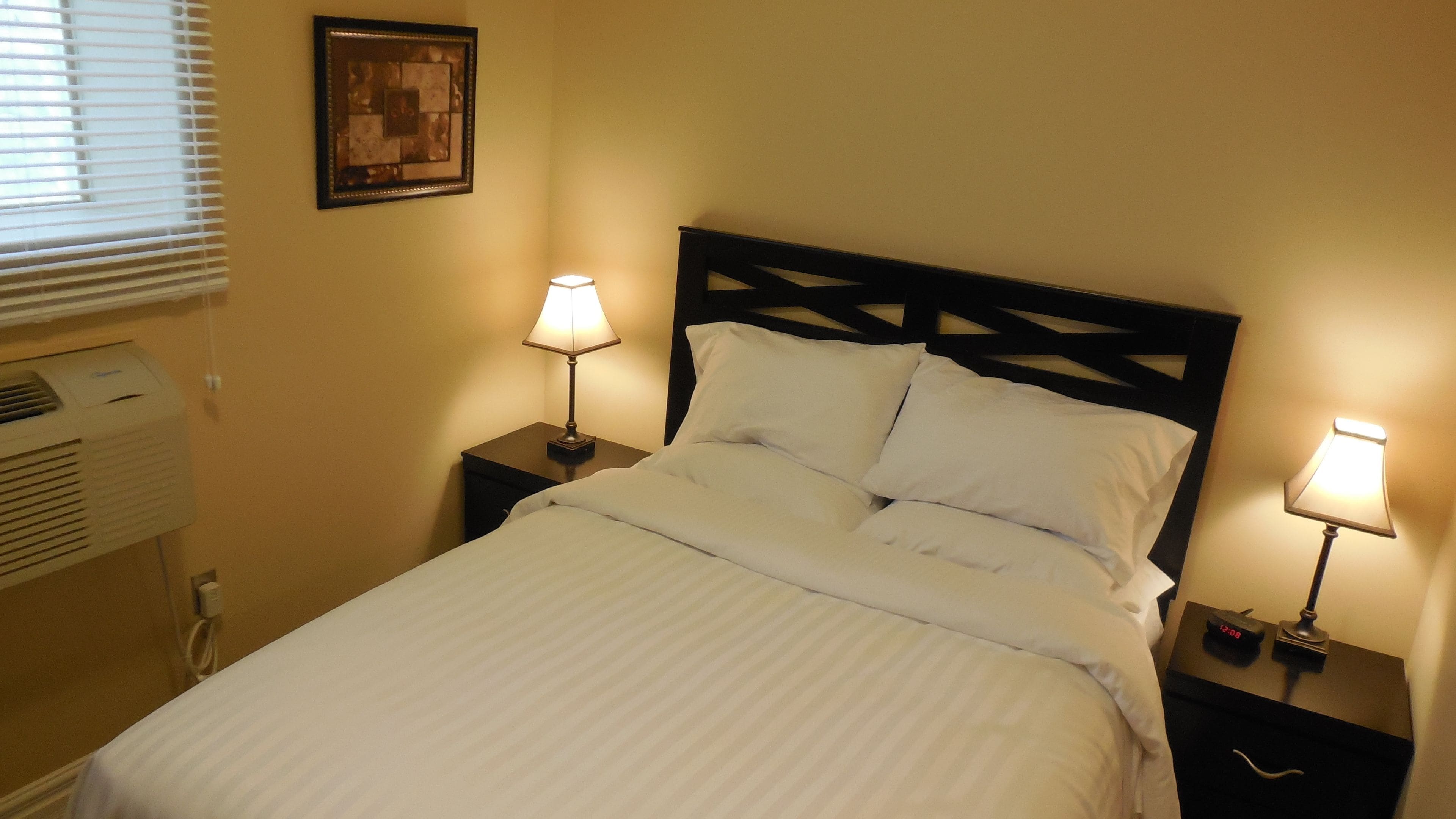 A neatly made bed with white linens, two bedside lamps, a wall-mounted air conditioner, and a framed picture above the bed.