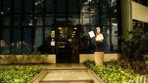 Entrada de un edificio con una estatua de un chef sosteniendo un menú. Hay escalones que conducen a la entrada, y un jardín con plantas verdes a ambos lados.