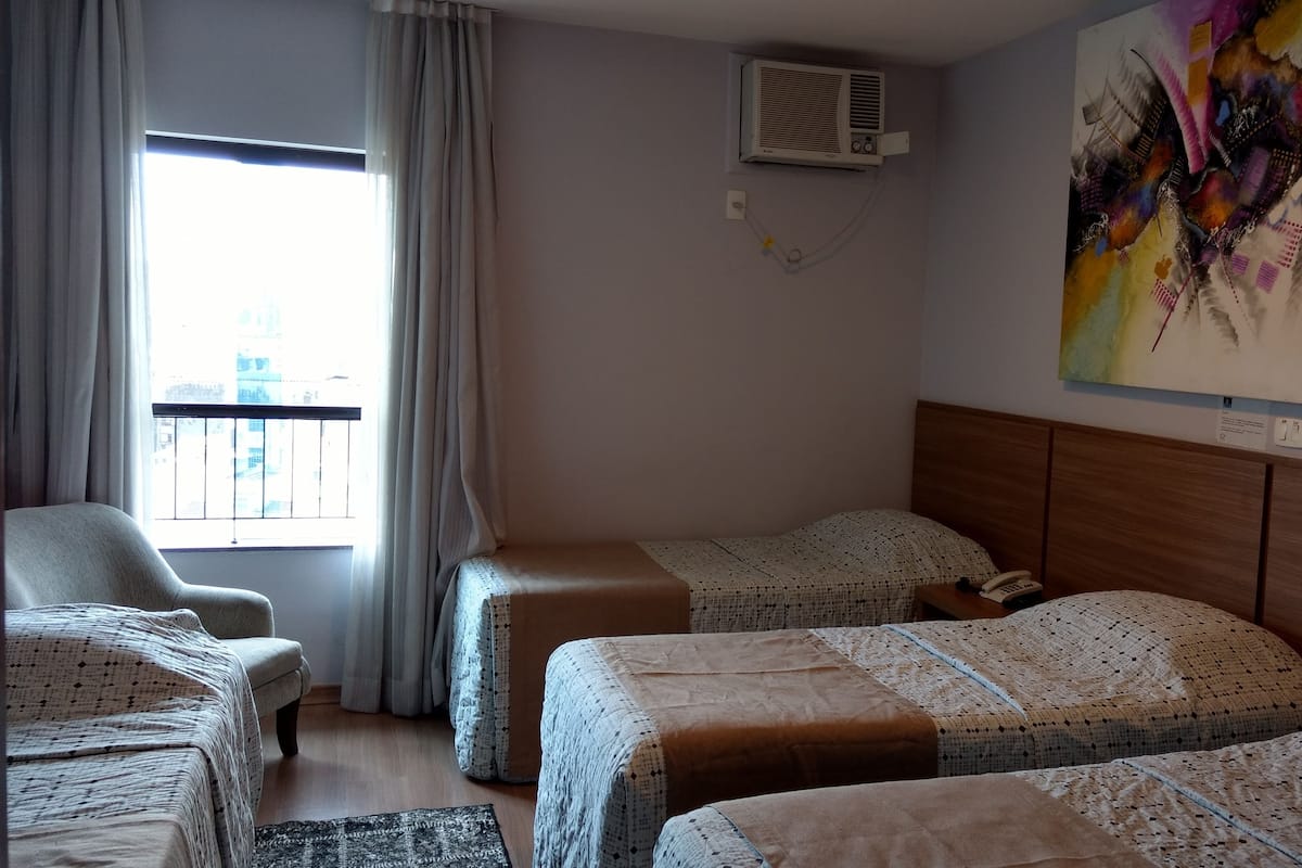 Une chambre d’hôtel avec deux lits simples, une chaise, une petite table, un appareil de climatisation et une fenêtre avec des rideaux.