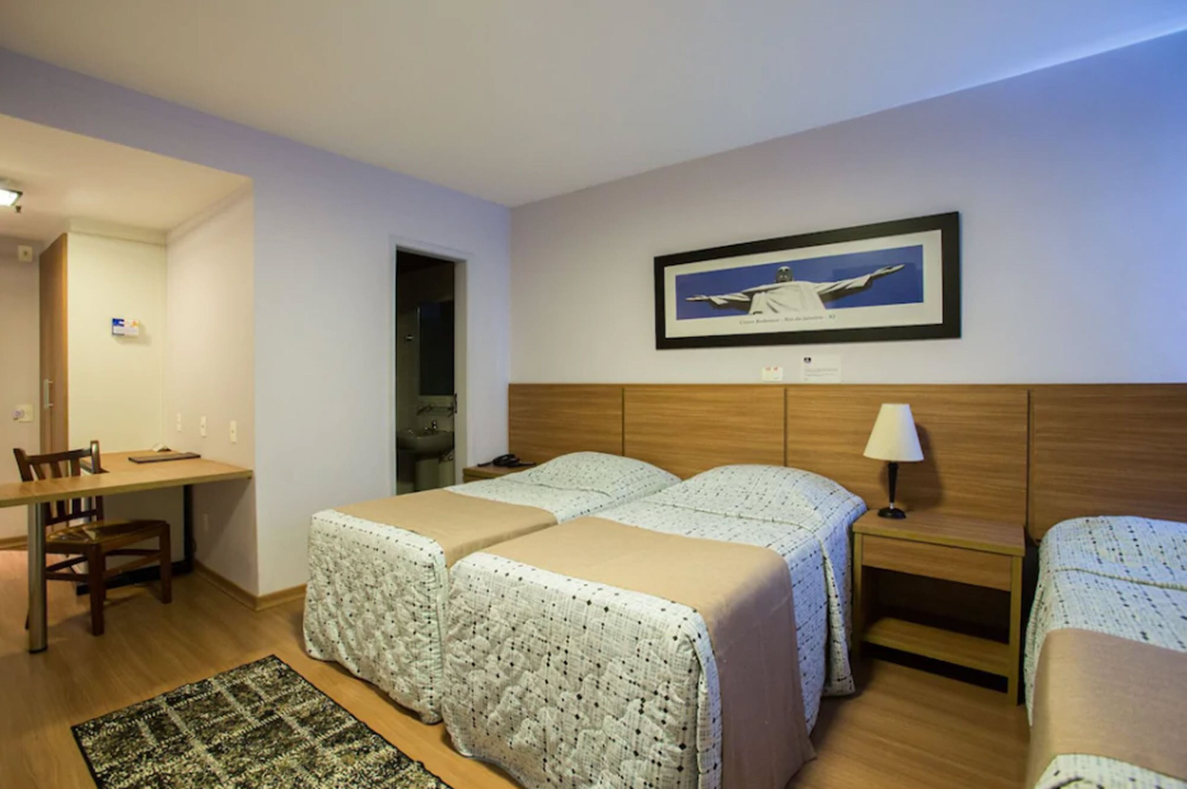 A hotel room with two beds, a desk, a chair, a lamp, and a framed picture on the wall.