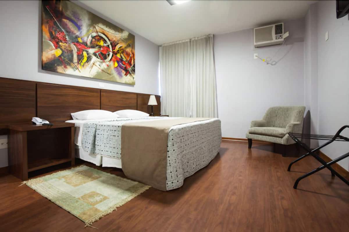 Une chambre d’hôtel avec un lit, une chaise, un bureau avec un téléphone, un tableau au mur et un appareil de climatisation.