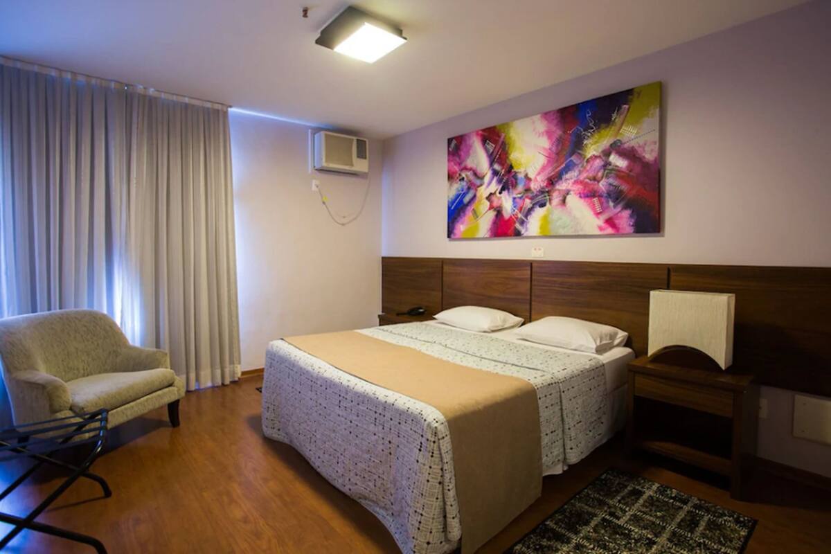 Une chambre d’hôtel avec un grand lit, une chaise, une table de chevet et une peinture abstraite aux couleurs vives accrochée au mur.