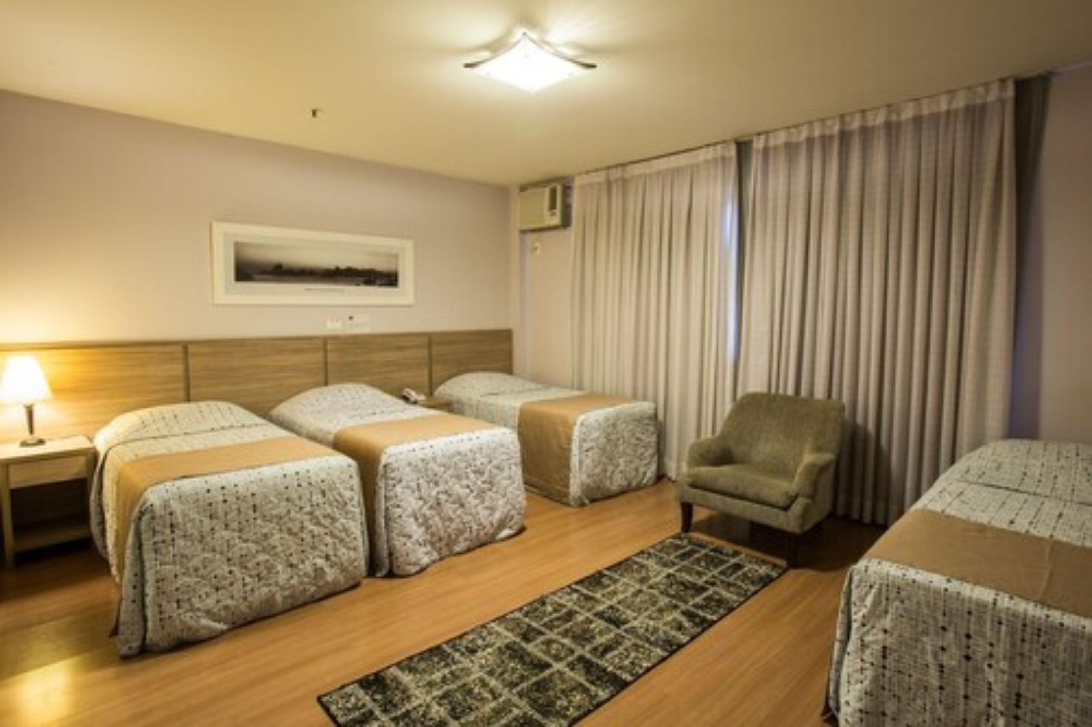 Une chambre d’hôtel avec deux lits, une chaise, une table de chevet, une lampe, un tableau encadré au mur et une fenêtre avec des rideaux.