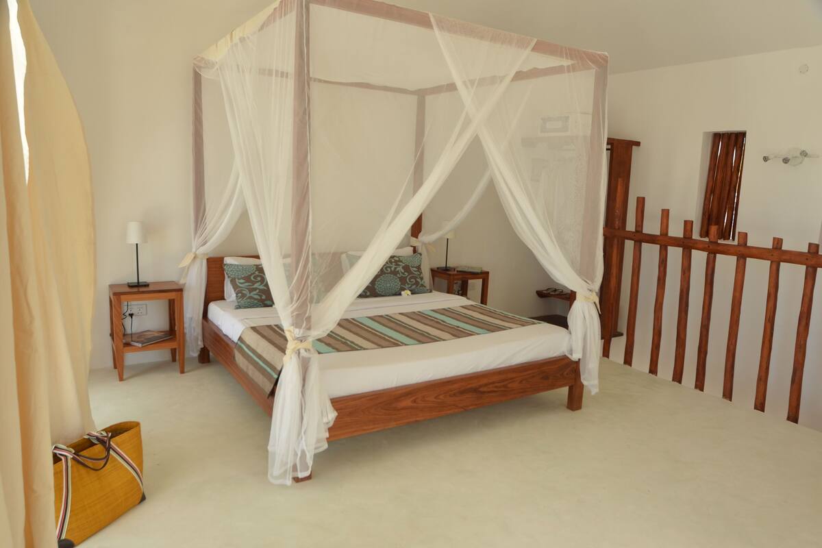 A four-poster canopy bed with a wooden frame and white bedding. There are two bedside tables with lamps and a small nightstand with a lamp. A wooden fence is visible in the background.