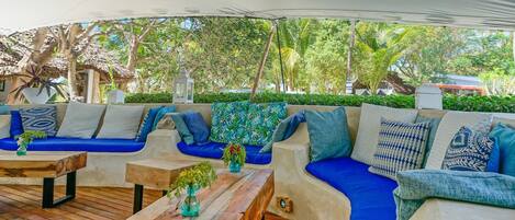 An outdoor seating area with blue cushioned sofas and wooden tables, surrounded by lush greenery and a thatched roof structure.