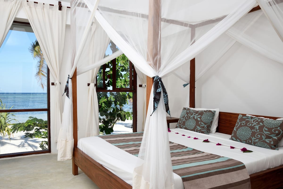 A bedroom with a four-poster canopy bed, a view of the ocean, and a tropical plant outside the window.