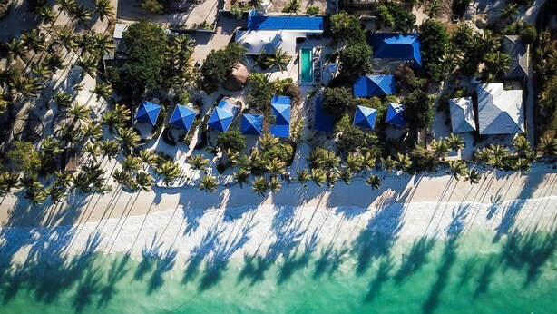 An aerial view of a resort with multiple blue-roofed buildings, a sandy beach, and palm trees. - Indigo Beach Zanzibar (Bwejuu)