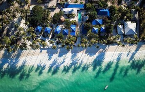 An aerial view of a resort with multiple blue-roofed buildings, a sandy beach, and palm trees. - Indigo Beach Zanzibar (Bwejuu)