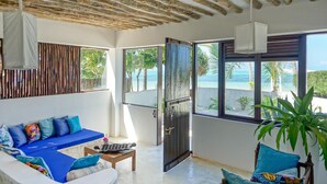 A room with a wooden ceiling, large windows with a view of the sea, a blue sofa with colorful cushions, and a potted plant. - Indigo Beach Zanzibar (Bwejuu)