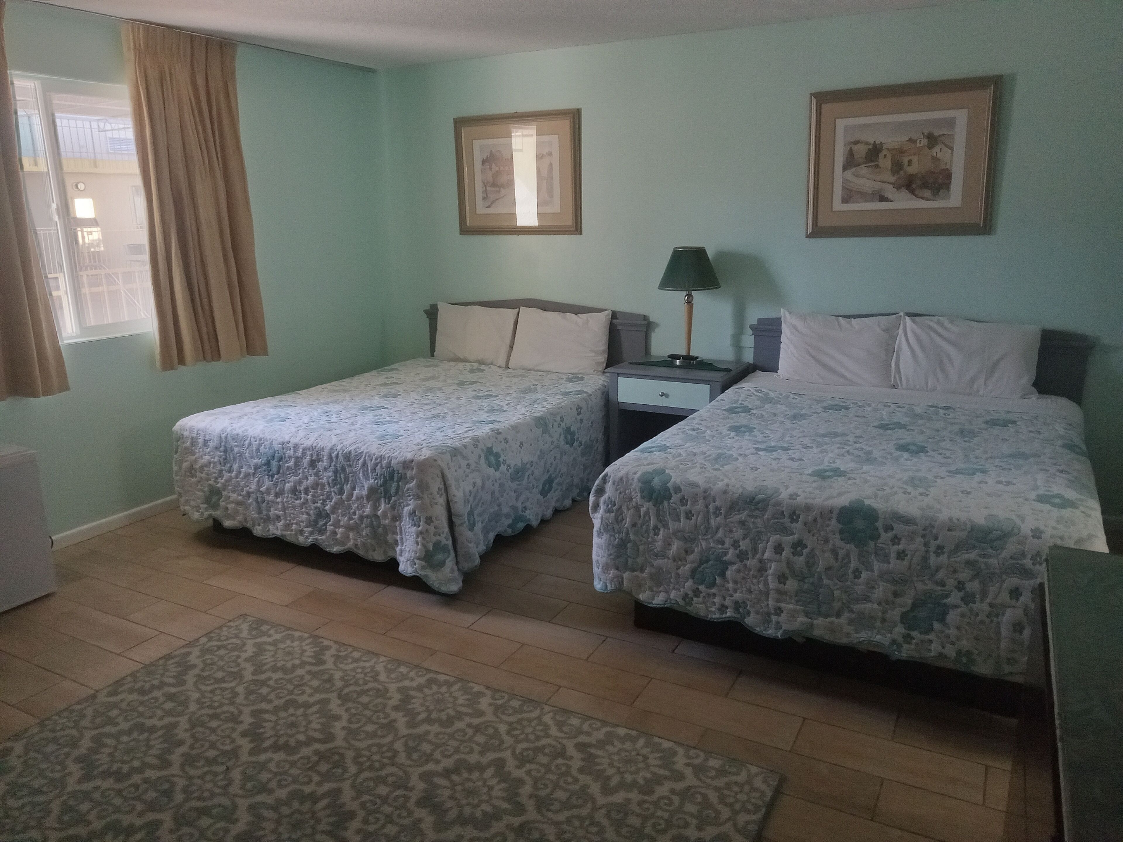 A room with two single beds, a nightstand with a lamp, a window with curtains, and two framed pictures on the wall.