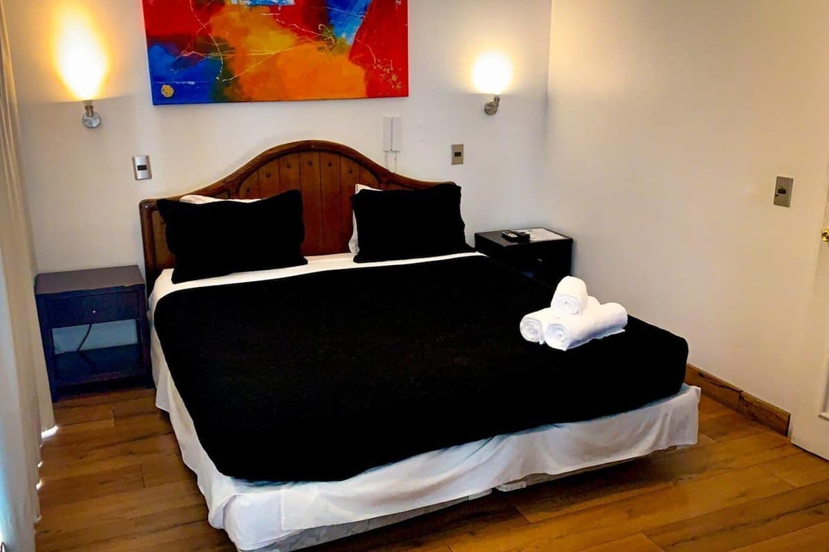 Un lit bien fait, avec une couvre-lit noire et des draps blancs, une tête de lit en bois, deux lampes de chevet et une petite table de chevet avec une télécommande.