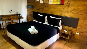 Une chambre d’hôtel avec une tête de lit en bois, un lit recouvert de draps blancs, deux serviettes enroulées posées sur le lit, une petite table de chevet en bois avec une télécommande, et un bureau en bois avec une lampe et un cadre photo.