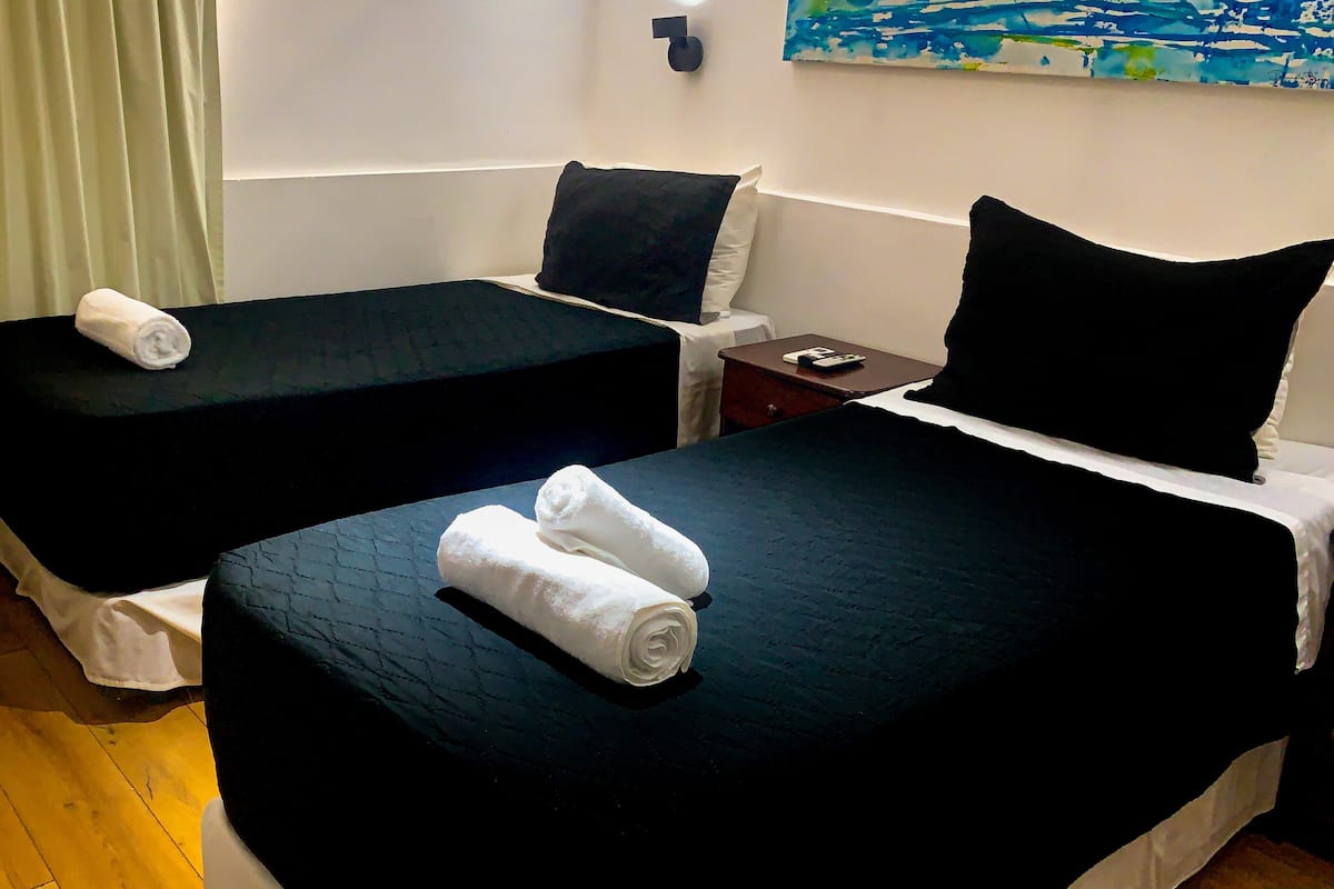 Une chambre d’hôtel avec deux lits, chacun doté de draps noirs et de serviettes blanches. On y trouve une petite table de chevet en bois, une lampe et un téléphone.