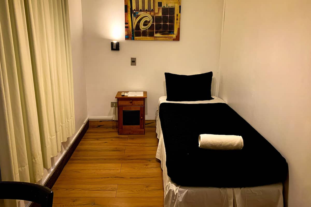 Une petite chambre avec un lit simple, un plancher en bois, une table de chevet, une lampe fixée au mur et un tableau abstrait aux couleurs vives.