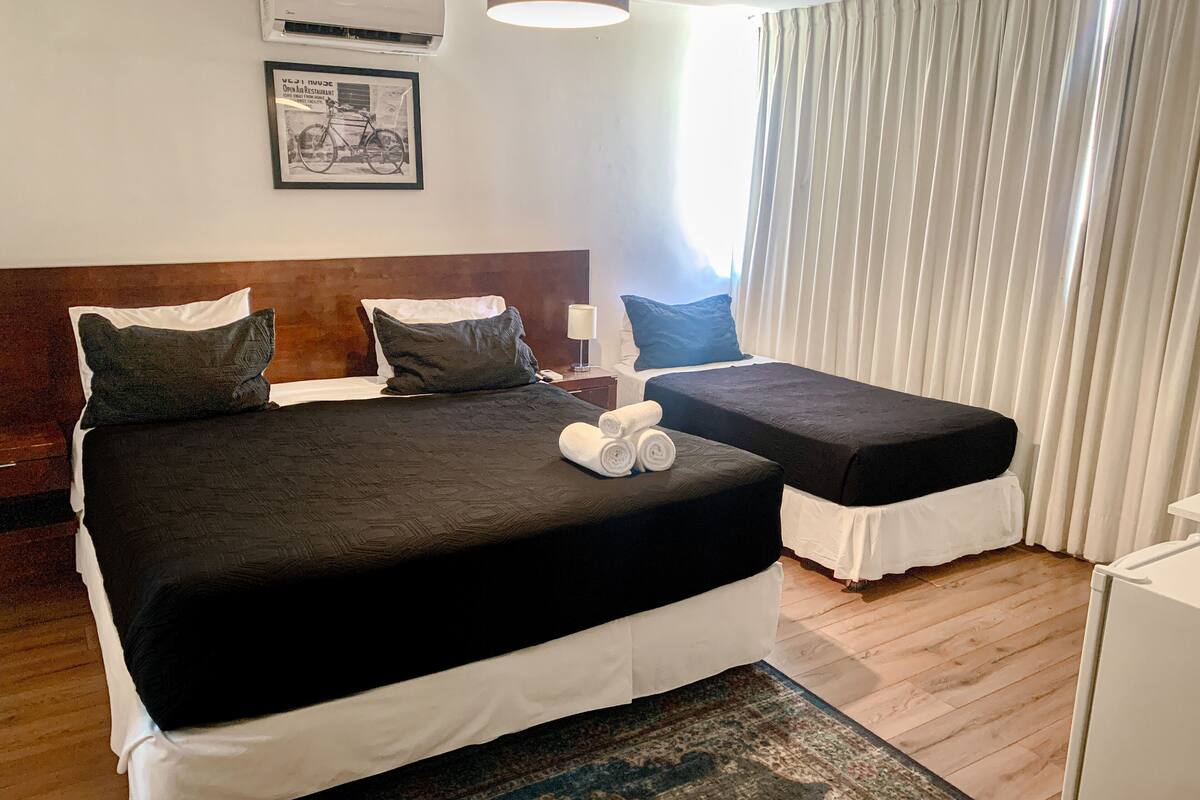 Un lit double avec une couvre-lit noire et des draps blancs, une petite table de chevet avec une lampe, et une fenêtre avec des rideaux blancs.