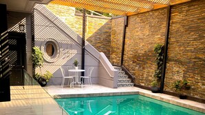 Un espace piscine intérieur moderne, doté d’une paroi en verre transparent, d’un escalier menant à un balcon et d’un coin salon avec des chaises et une table.