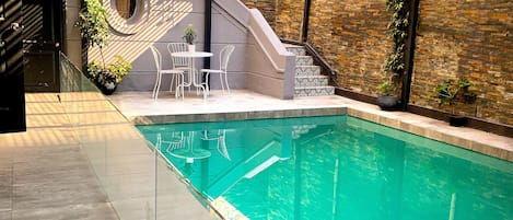 A modern indoor pool area with a clear glass barrier, a staircase leading to a balcony, and a seating area with chairs and a table.
