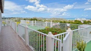 A balcony with white railings, a paved walkway, and a view of a coastal area with buildings and a cruise ship in the distance.