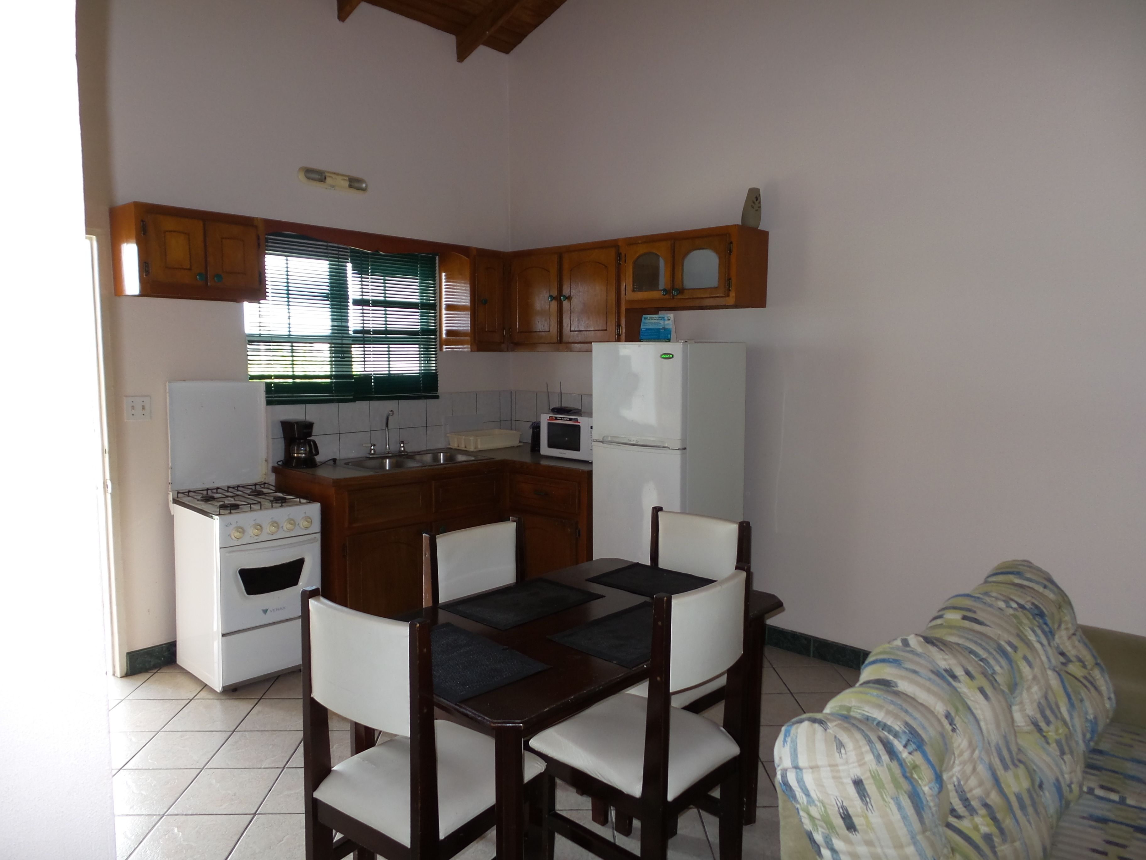 A compact kitchen with wooden cabinets, a gas stove, a microwave, and a refrigerator. There is a dining table with chairs and a patterned sofa.