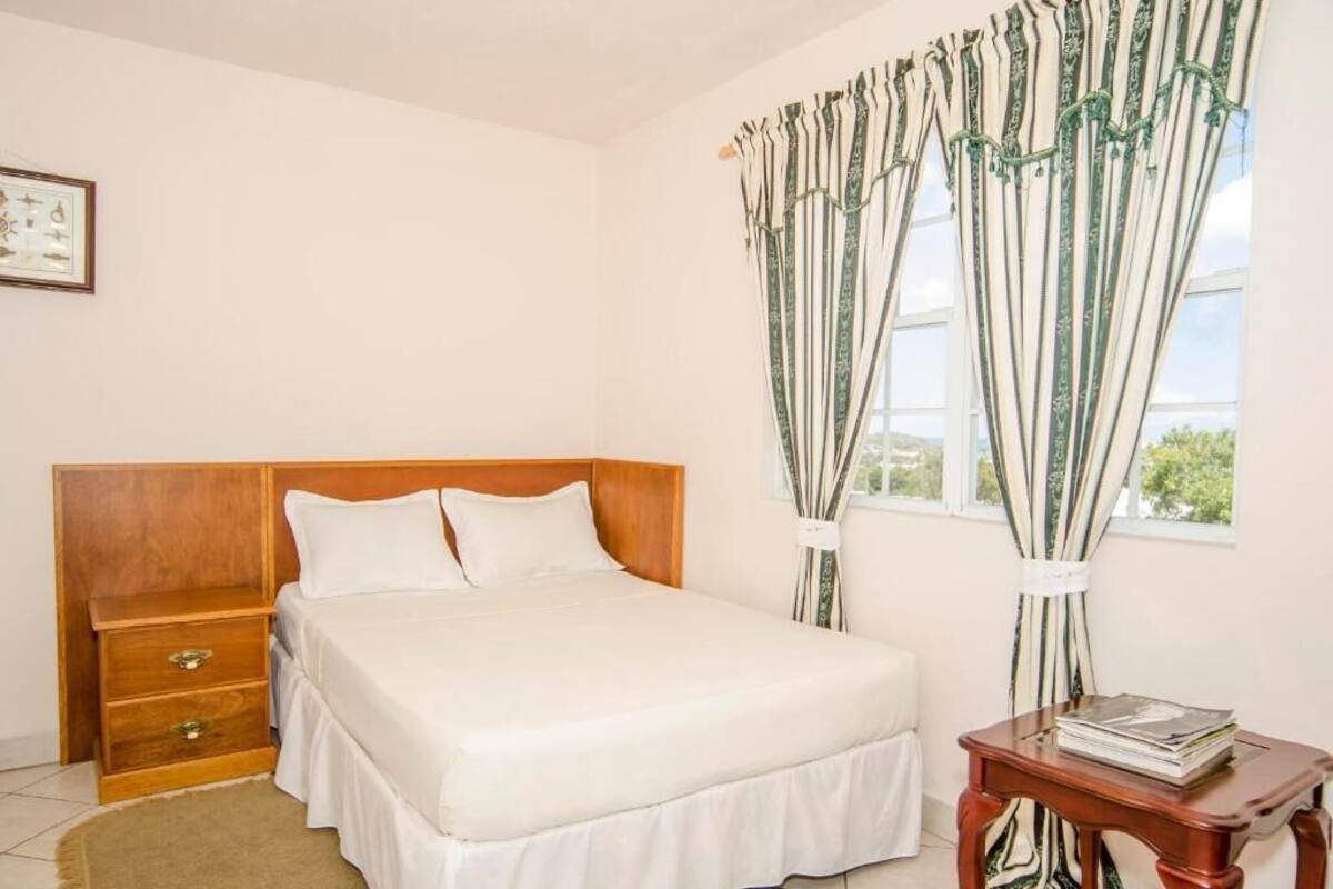 A neatly made bed in a room with a wooden headboard, a bedside table with a book, and curtains on the window.