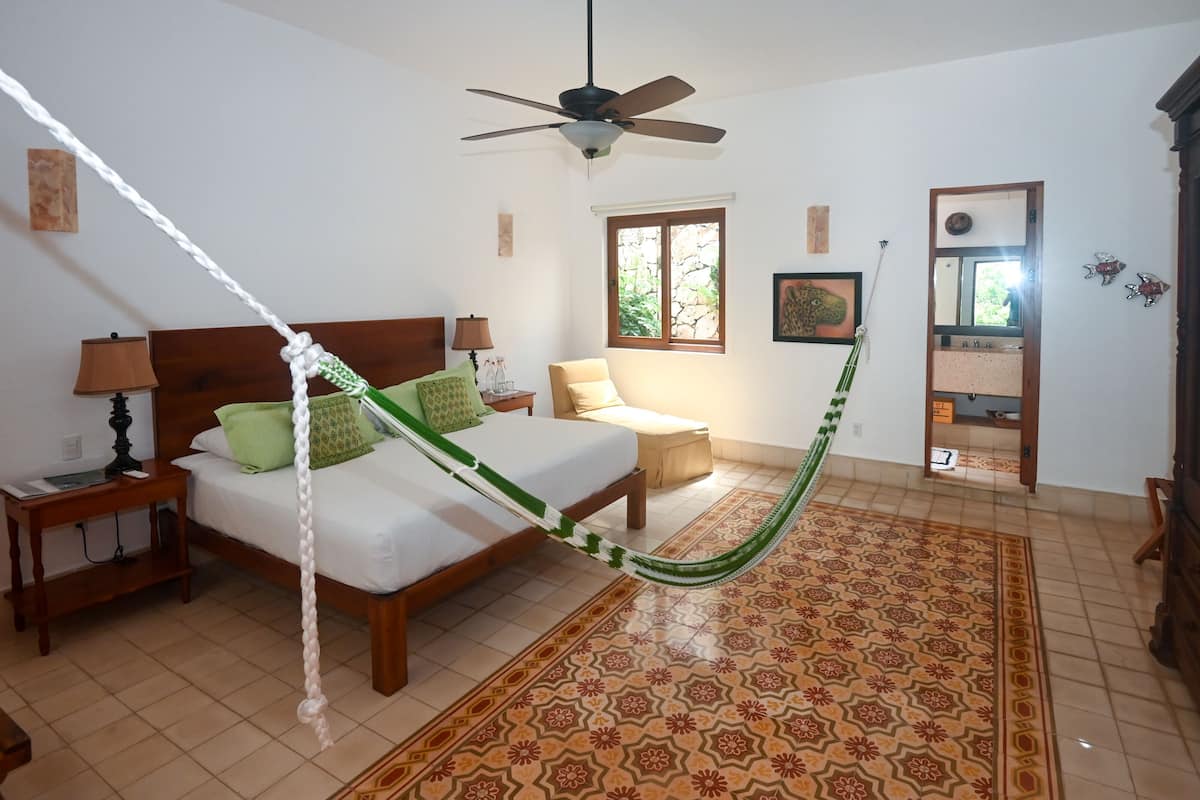 Quarto com cama, mesa de cabeceira, luminária, cadeira, rede, ventilador de teto, janela e espelho.