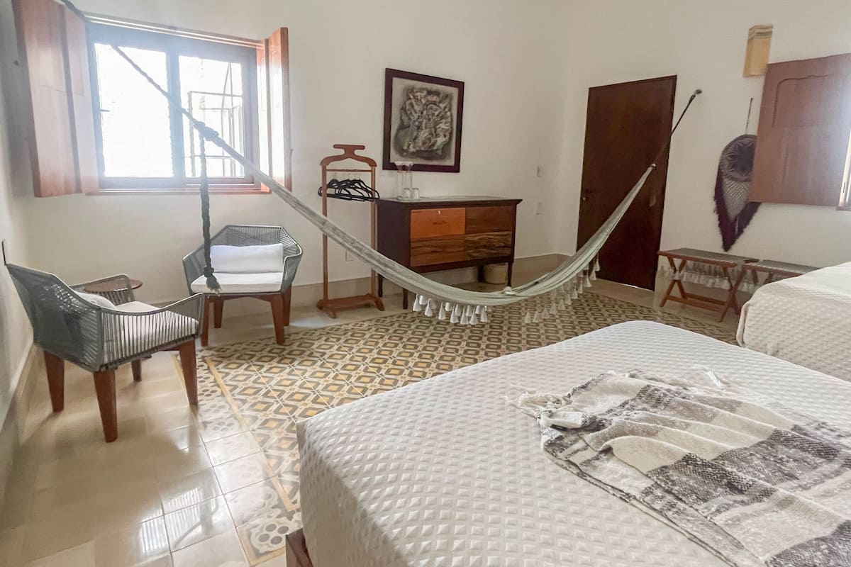 Quarto de hotel com cama, cadeira, escrivaninha, cadeira suspensa, cómoda e janela.