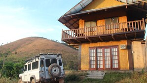 A traditional wooden house with a balcony, a vehicle parked in front, and a hill in the background.