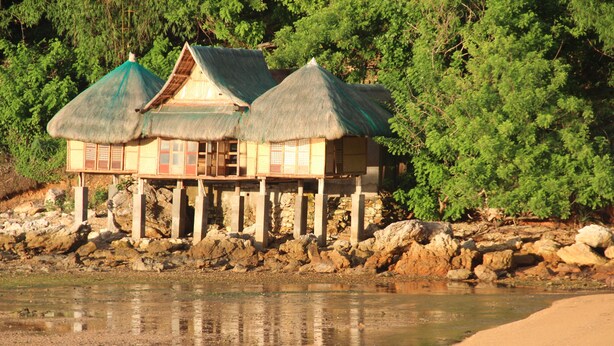 A traditional wooden house with a thatched roof, situated on stilts over a body of water, surrounded by dense greenery.