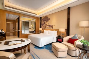Una habitación de hotel moderna con una cama grande, una zona de estar con sofá y reposapiés, un escritorio con lámpara y una mesa de comedor.