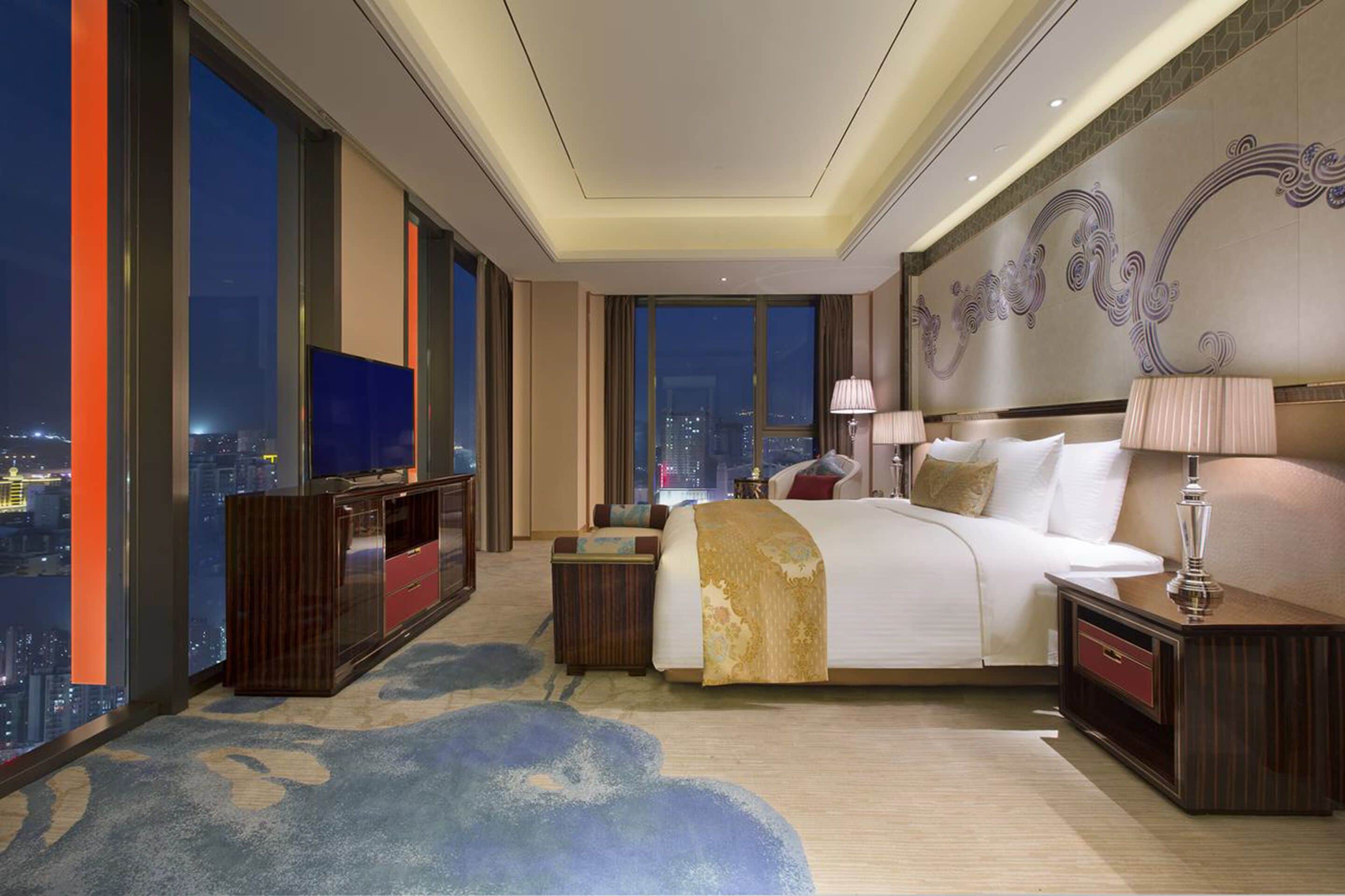 A spacious hotel room with a large bed, a TV, a desk, and a view of the city at night.