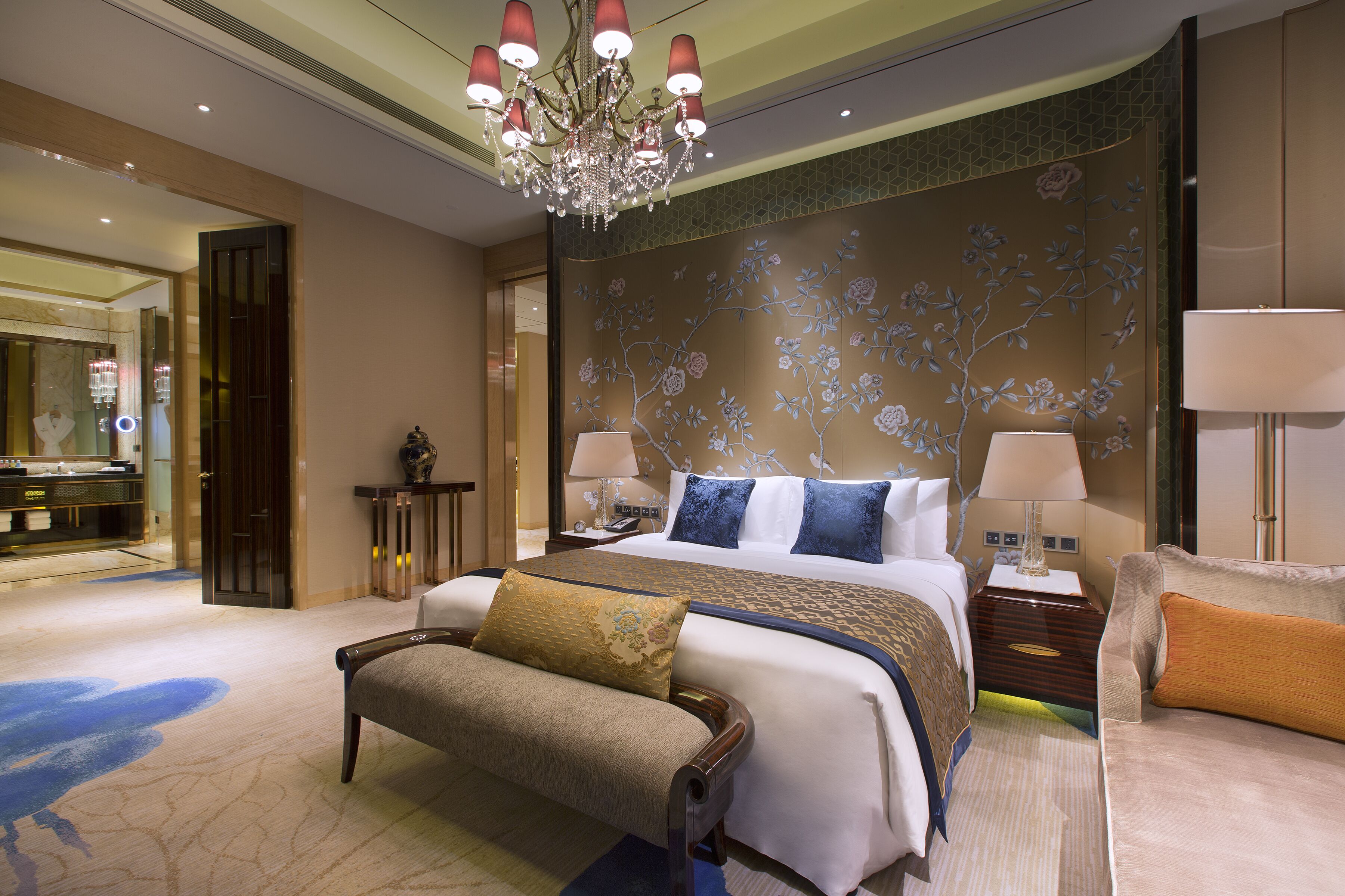Une chambre d’hôtel avec un grand lit, deux tables de chevet, un banc, un lustre et un mur décoratif.