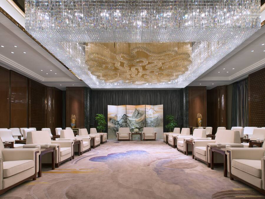 A spacious conference room with a large, ornate chandelier, multiple seating arrangements, and a large painting on the back wall.