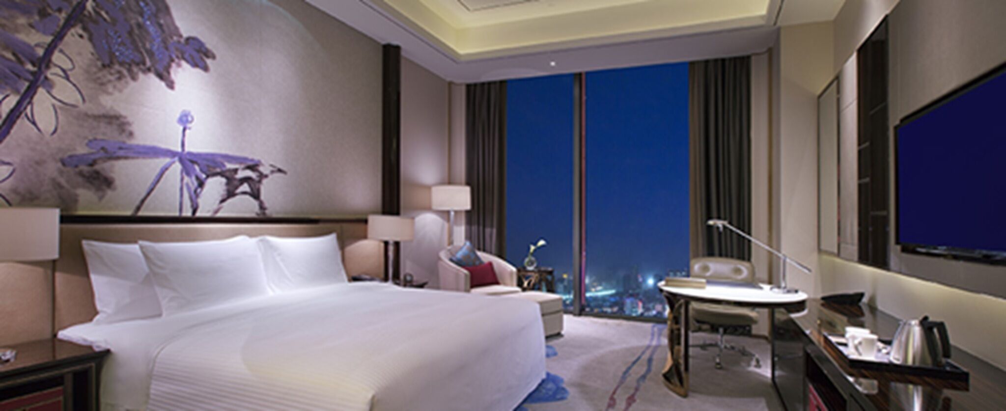 A hotel room with a large bed, a desk, a chair, a small table, and a large window with a city view.
