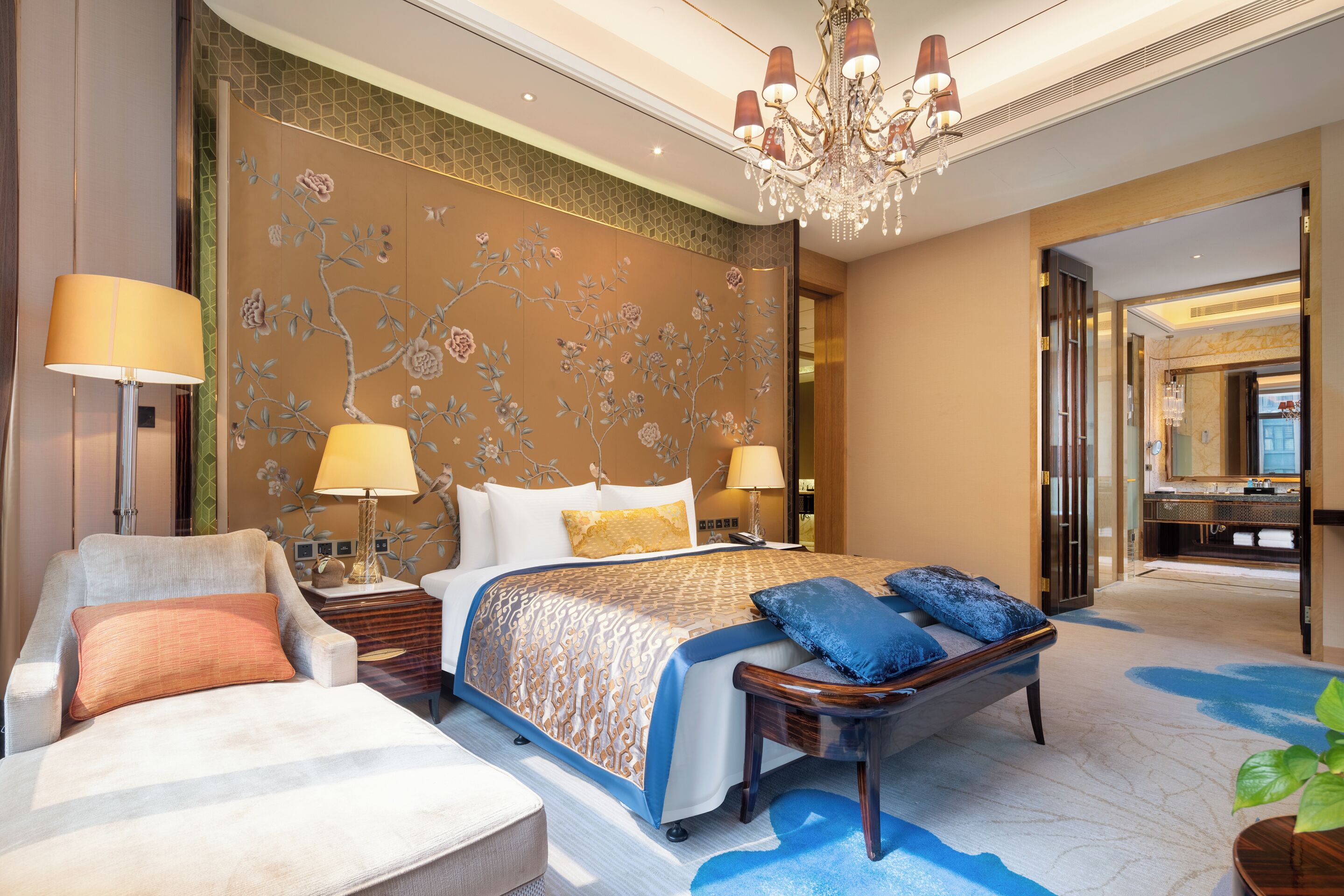 A well-appointed hotel room with a large bed, a sitting area with a chair and a small table, a decorative wall, and a chandelier.