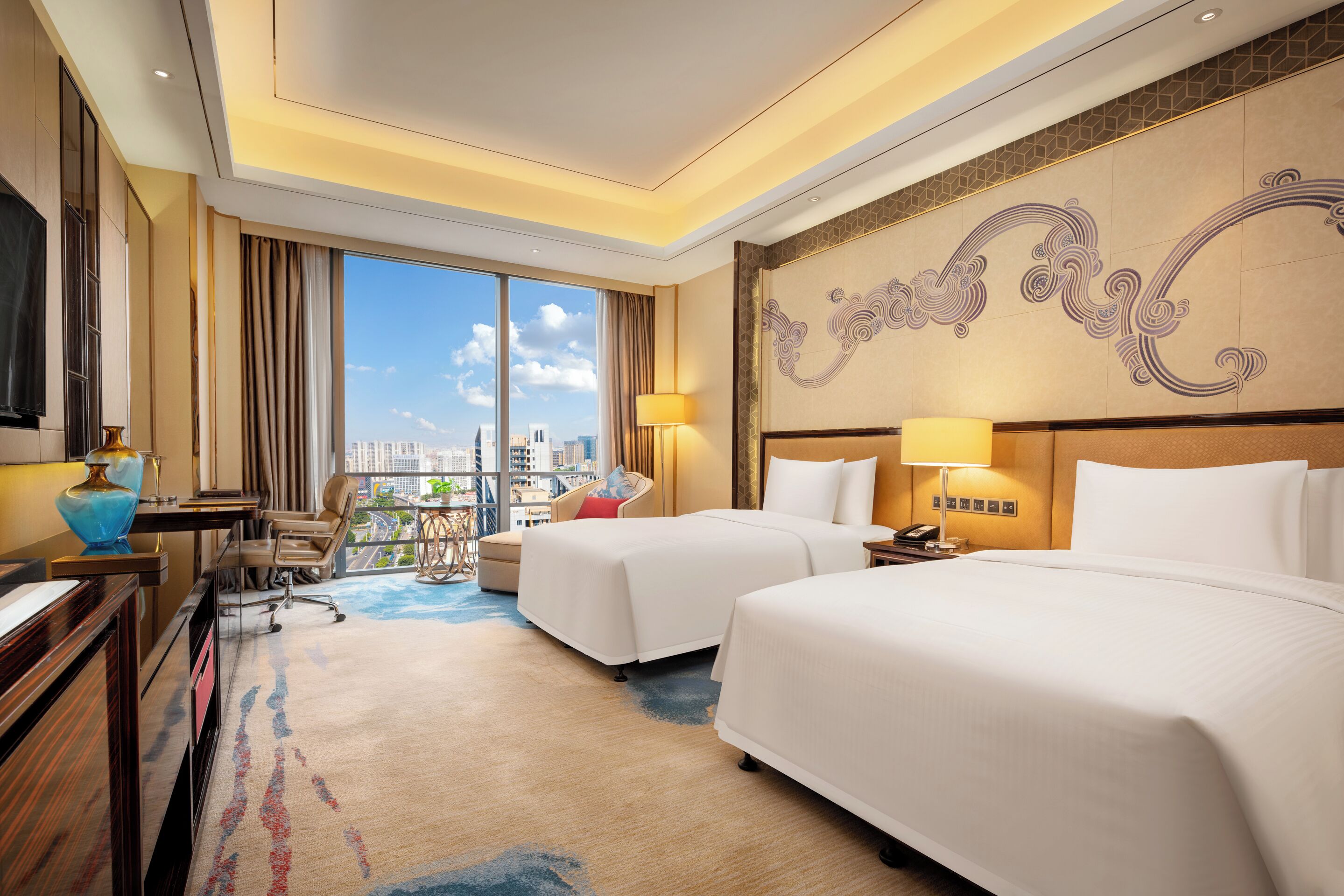 A hotel room with two beds, a desk, a chair, a TV, and a balcony with a city view.
