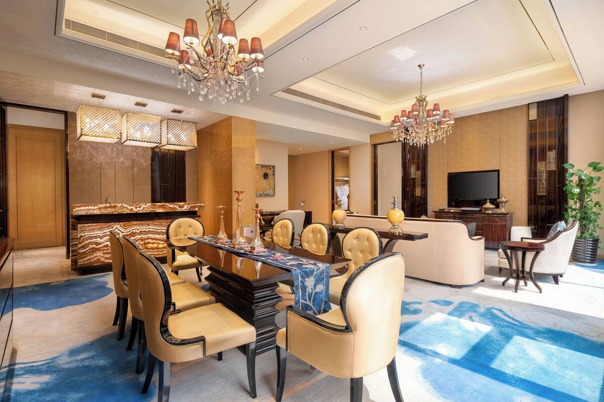 Een modern restaurantgedeelte van een hotel, met een grote tafel, stoelen, kroonluchters en een televisie.