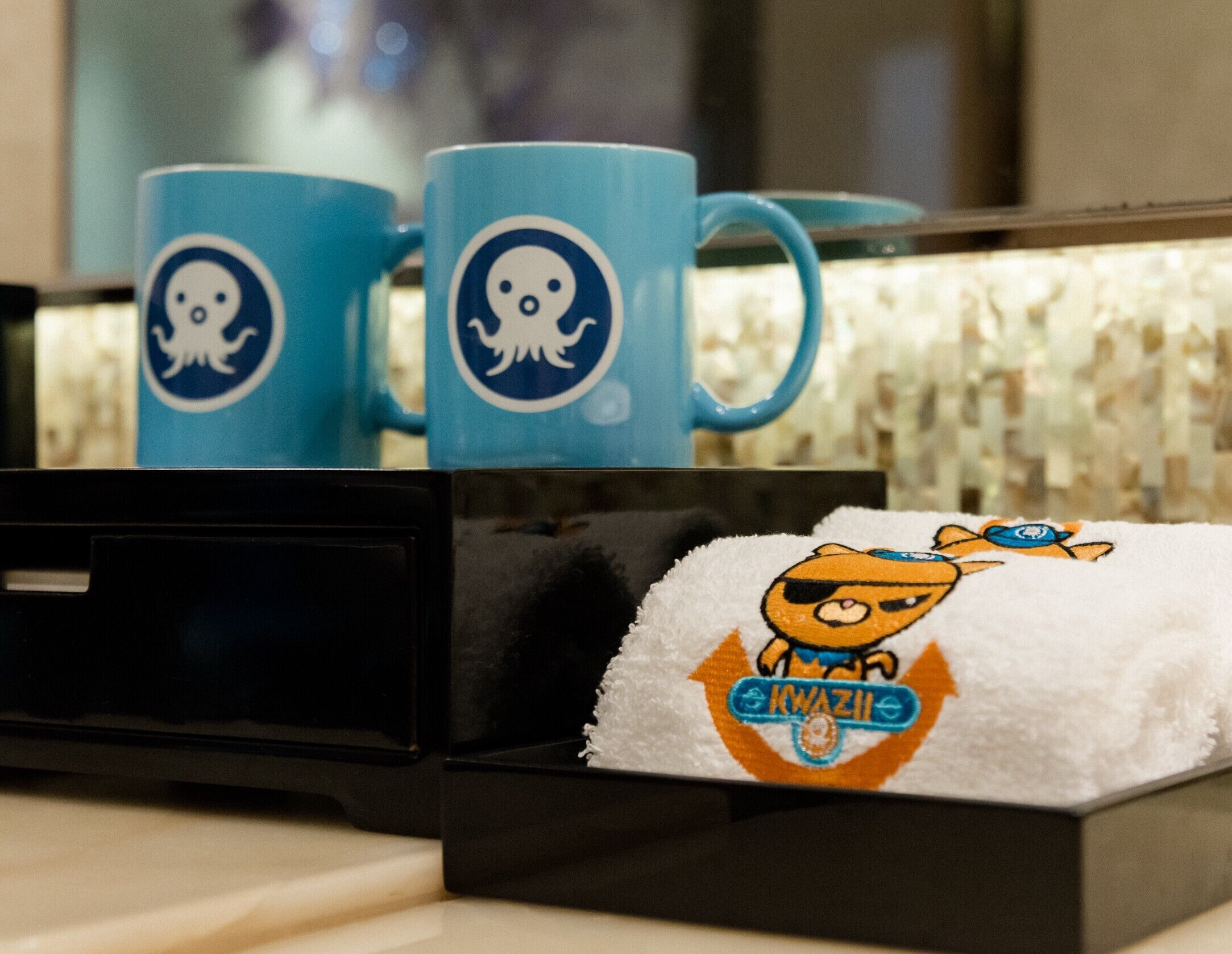 A hotel room with a minibar containing two blue mugs with a white octopus logo, a towel with a cartoon character, and a small box of white fluffy material.