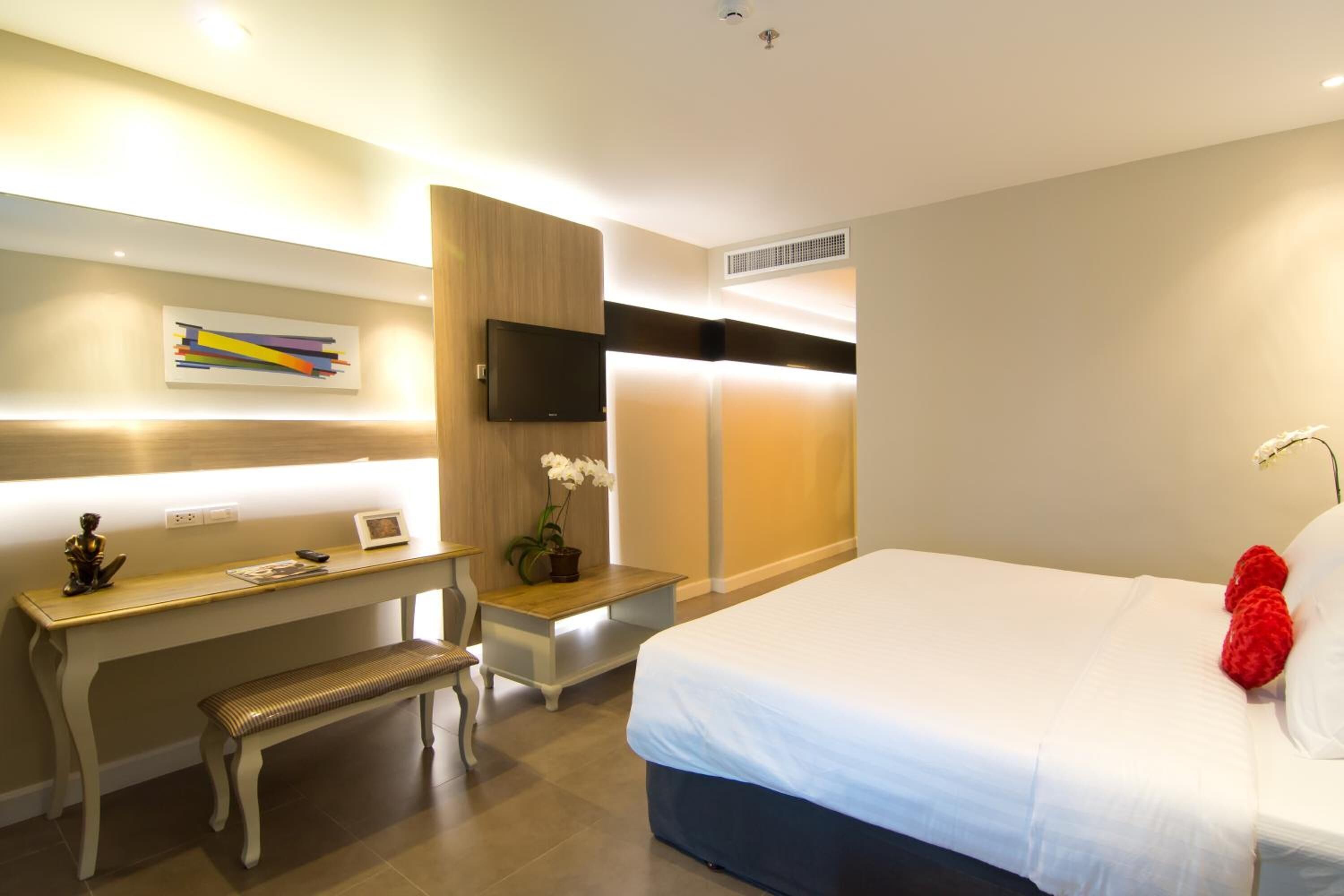 Une chambre d’hôtel comprenant un lit, un bureau, une banquette, une télévision et une étagère murale ornée d’un tableau.