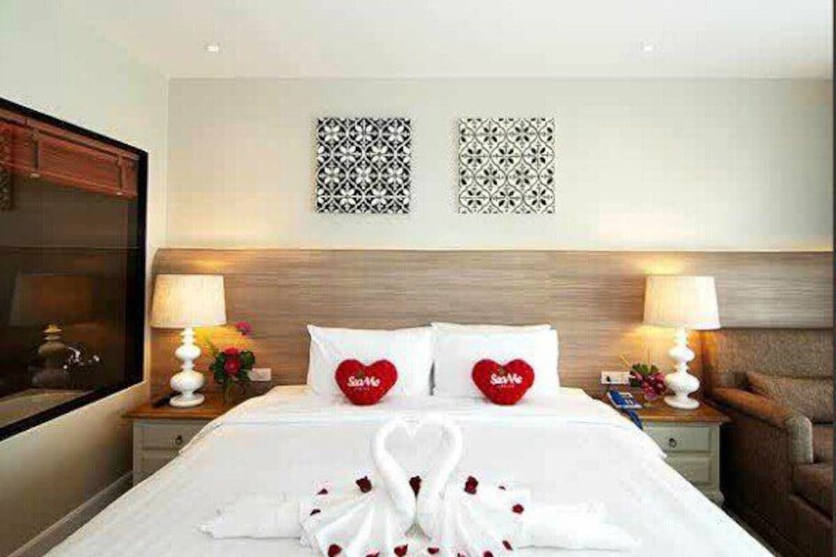 A hotel room with a neatly made bed, two heart-shaped pillows, and a decorative wall piece above the bed.