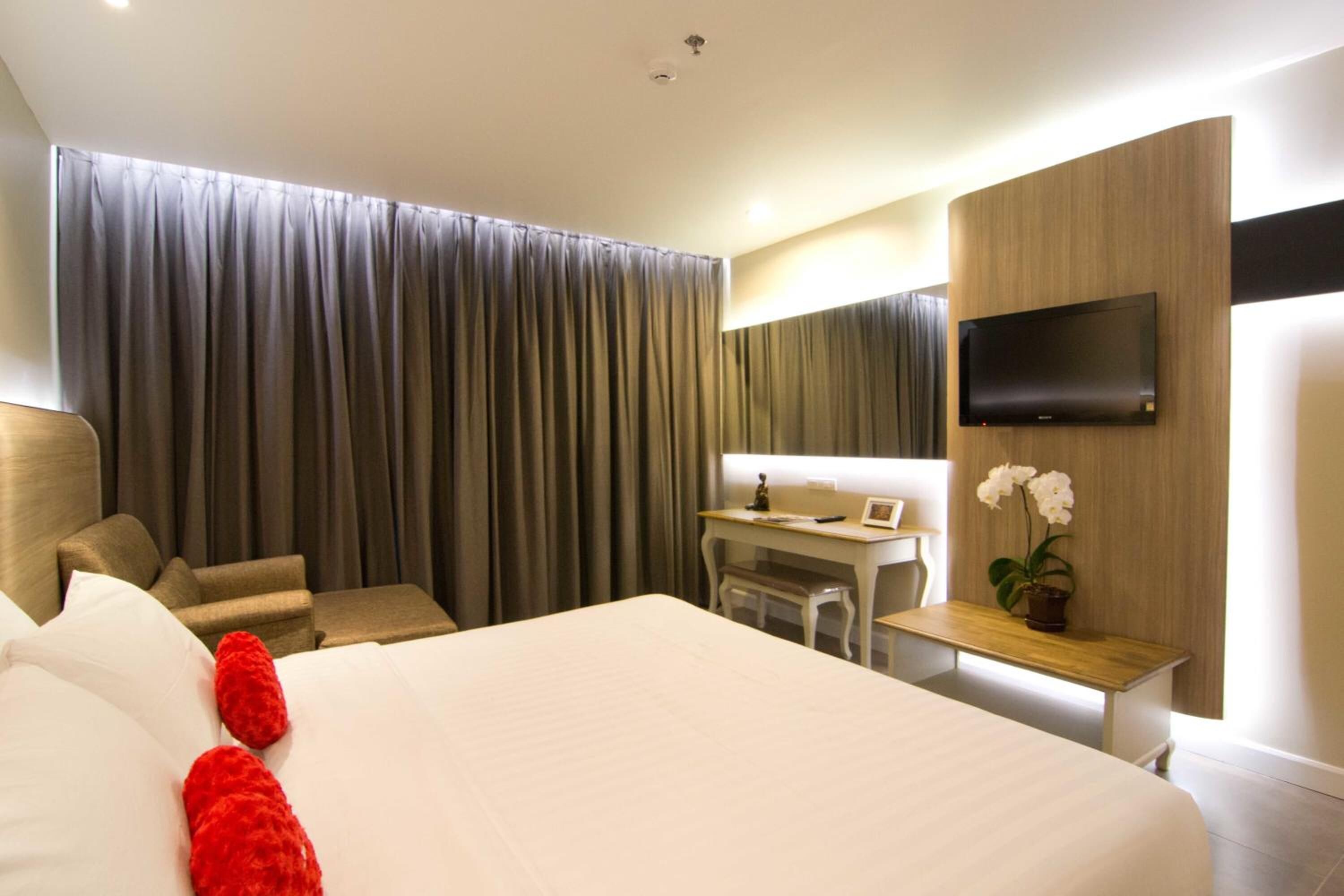 A hotel room with a large bed, a desk, a chair, a TV mounted on the wall, and a window with curtains.
