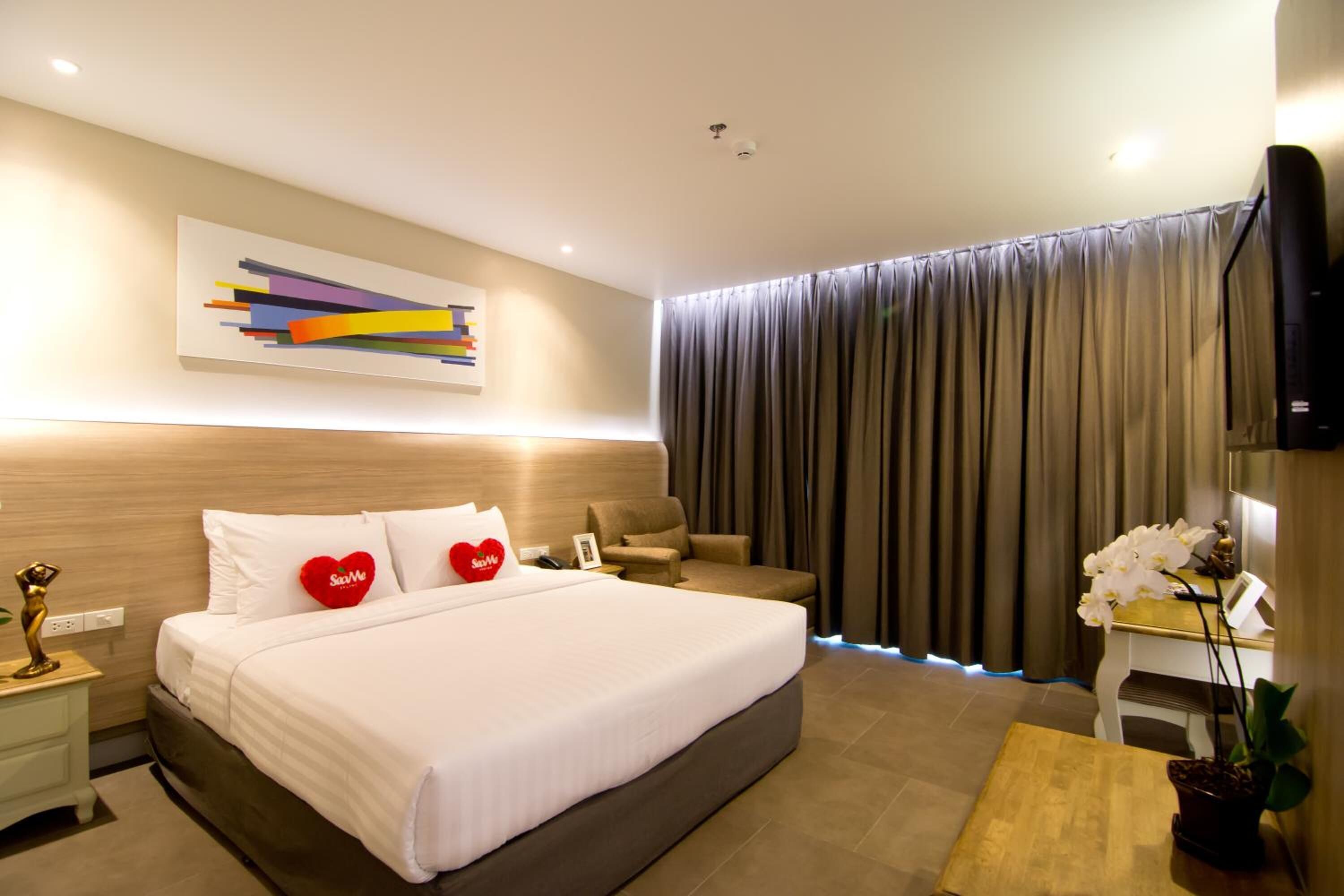 A modern hotel room with a large bed, a bedside table with a lamp, a small desk, and a television.