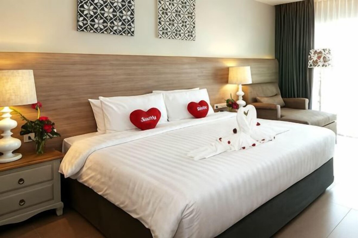 A neatly made bed with white linens and heart-shaped pillows, a bedside table with a lamp, and a chair.